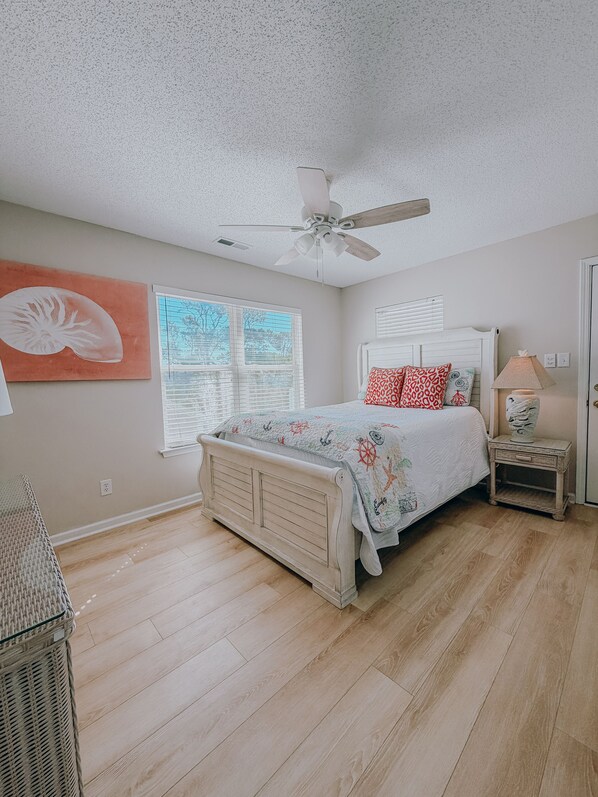 A bedroom with a bed, a ceiling fan, a window with blinds, a nightstand with a lamp, and a wall with a decorative piece. - NEWLY Furnished 6BR, Lake Views, Steps to Beach, Pool & Amenities (Myrtle Beach)