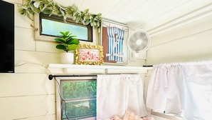A cozy room with a bed, a window with curtains, a wall-mounted TV, a fan, and a shelf with decorative items.