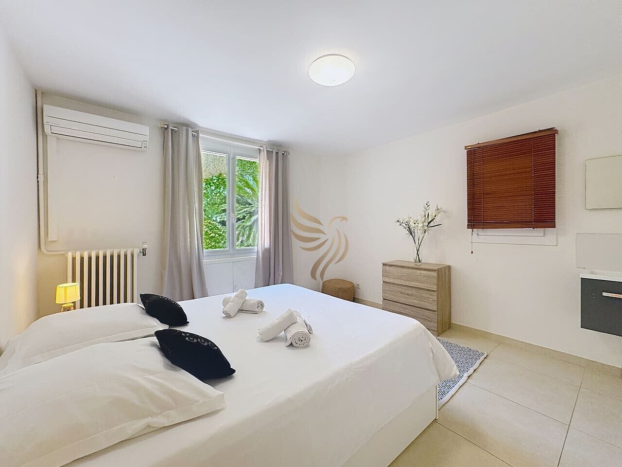 A hotel room with a large bed, a radiator, a window with curtains, a wooden dresser, a vase with flowers, and a wall-mounted TV.