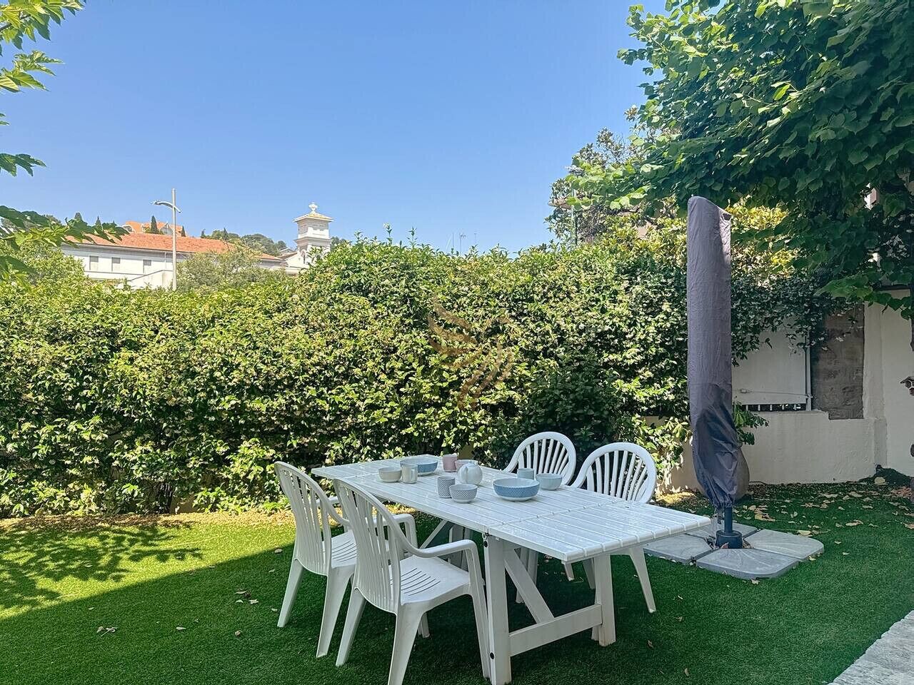 Un espace extérieur aménagé pour les repas, avec une table et des chaises blanches, entouré de pelouse verdoyante et d’une haie.