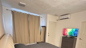 Un dormitorio con cama, cortinas, cómoda, televisor y aire acondicionado.