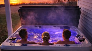 Un jacuzzi con tres personas sentadas adentro, rodeado por una terraza de madera y vistas a un atardecer.