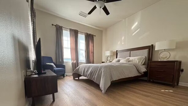 A bedroom with a bed, a desk, a chair, a ceiling fan, and a window with curtains. - 3 Bedrooms Condos at Cottages Oakleaf (Orange Park)