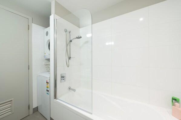 A modern bathroom with a glass-enclosed shower, a white bathtub, and a built-in washing machine. - Paihia Waterfront Suites (Paihia)