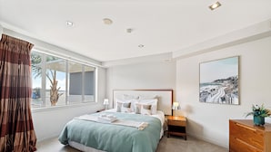 Quarto com cama grande, mesa de cabeceira, penteadeira, janela com cortinas e um quadro na parede.