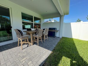 An outdoor patio with a wooden table and chairs, a grill, and a view of a residential area.