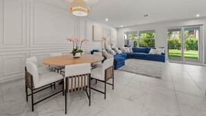 A modern living room with a dining area, featuring a round wooden table, white chairs, a blue sectional sofa, and a large window offering a view of a green garden.