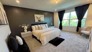 A bedroom with a large bed, a nightstand, a chair, a window with curtains, and a wall-mounted artwork.