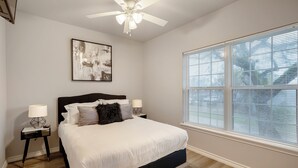 A bedroom with a large bed, a ceiling fan, a window with blinds, and a wall-mounted artwork.