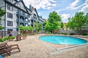 A swimming pool surrounded by a brick patio with wooden furniture and a stone wall.
