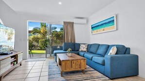 A modern living room with a blue sofa, a wooden coffee table, and a TV on a wooden stand. - Beachside Townhouse (Sunrise Beach)