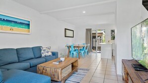A modern living room with a sofa, coffee table, and blue chairs. There is a TV on a wooden stand and a painting on the wall. - Beachside Townhouse (Sunrise Beach)