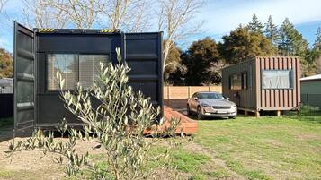A modern RV with a black exterior and a windowed cabin, parked in a grassy yard with a car and a small shed in the background.