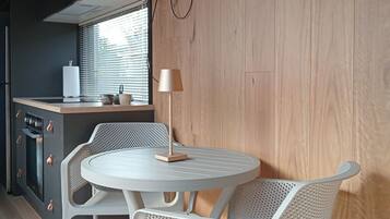 A modern kitchen with a built-in stove, a sink, and a window with blinds. There is a round dining table with two white chairs and a lamp. A flat-screen TV is mounted on the wall above the table.