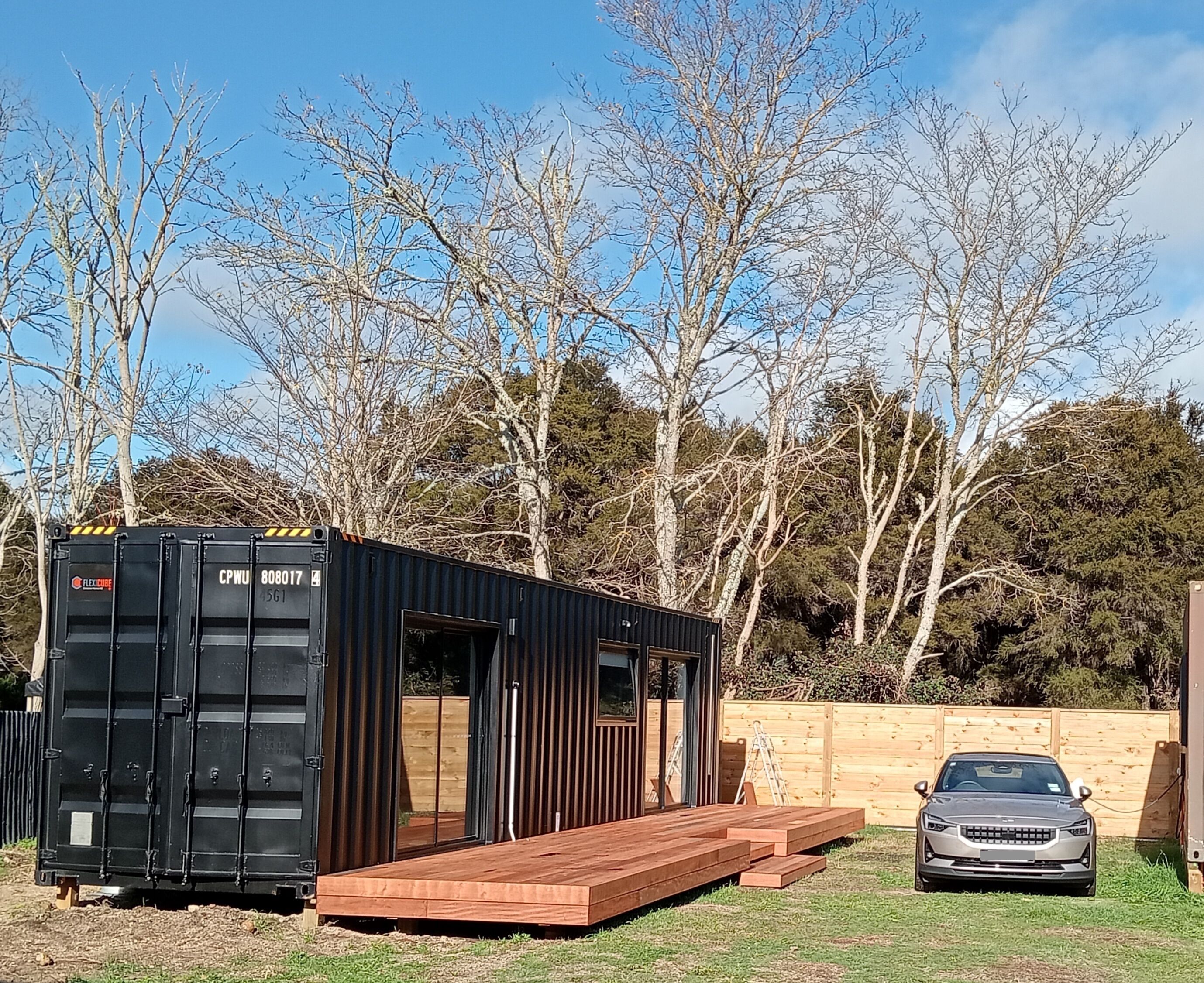 Una vivienda modular negra tipo contenedor, con una terraza de madera y un coche aparcado cerca.