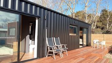 A modern, compact building with large sliding glass doors, a wooden deck with outdoor seating, and a clear view of the surrounding trees.