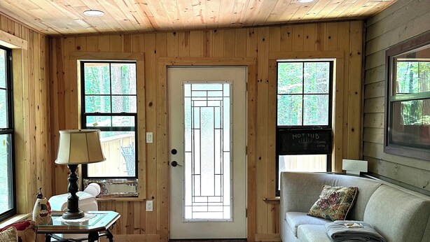 A cozy living room with wooden walls and floors, a sofa, a table with a lamp, and a door with a window. - Peaceful cozy cabin in the woods (Murphy)