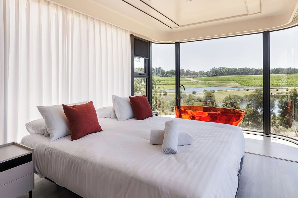 A modern bedroom with a large bed, white bedding, and a view of a lake and greenery.