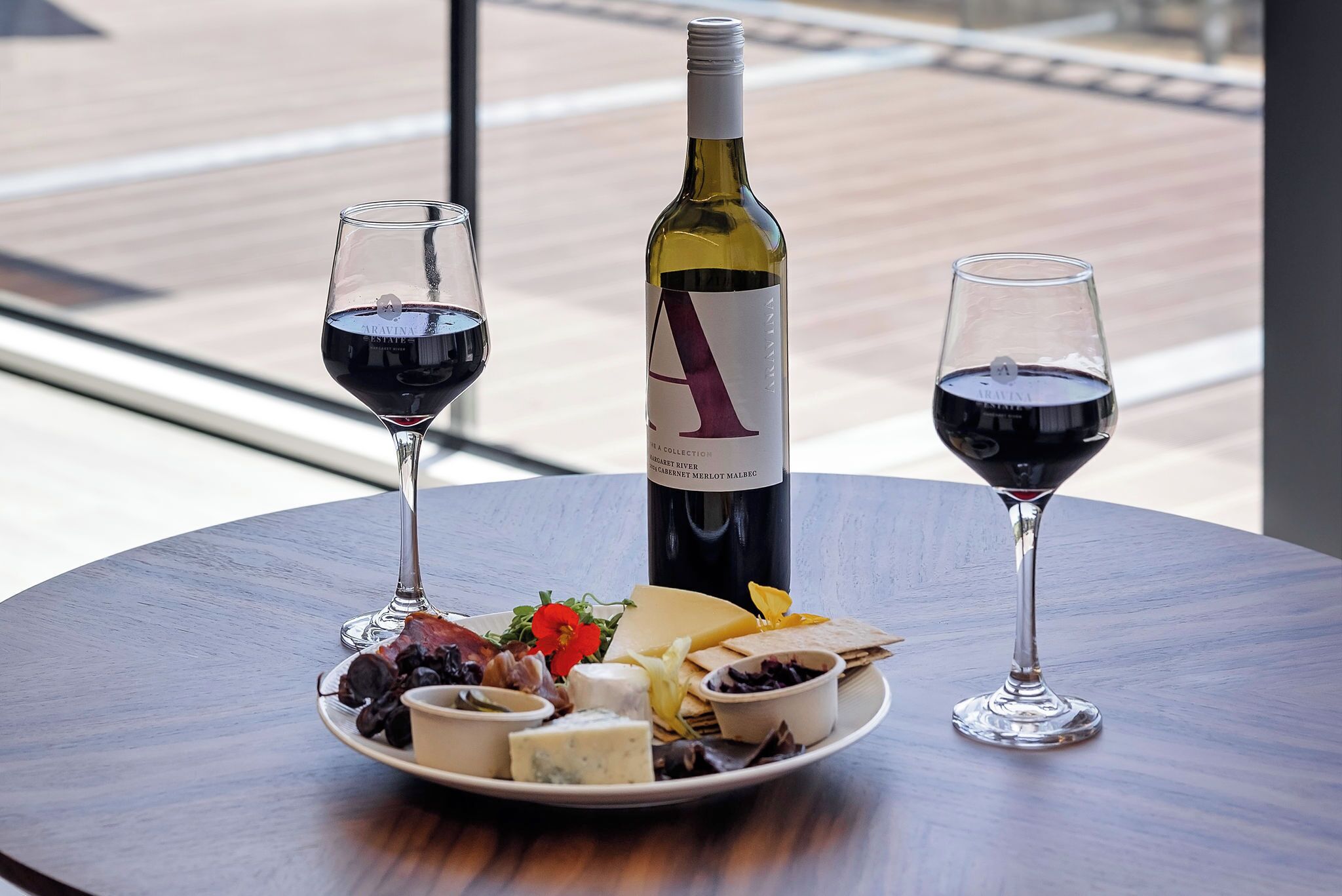 A table with a bottle of wine, two glasses of red wine, and a cheese platter with various accompaniments.