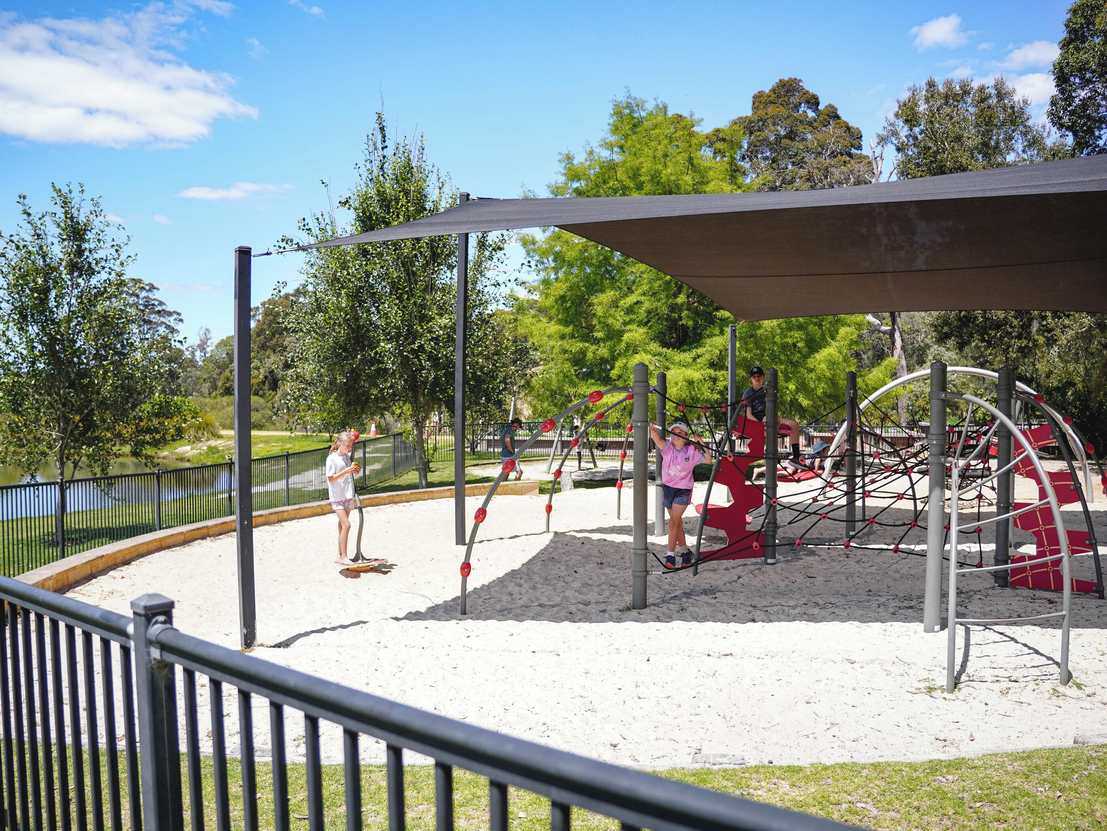 Un parque infantil con suelo de arena, juegos metálicos y una zona de descanso cubierta.