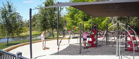 A playground with a sandy surface, metal play structures, and a covered seating area.