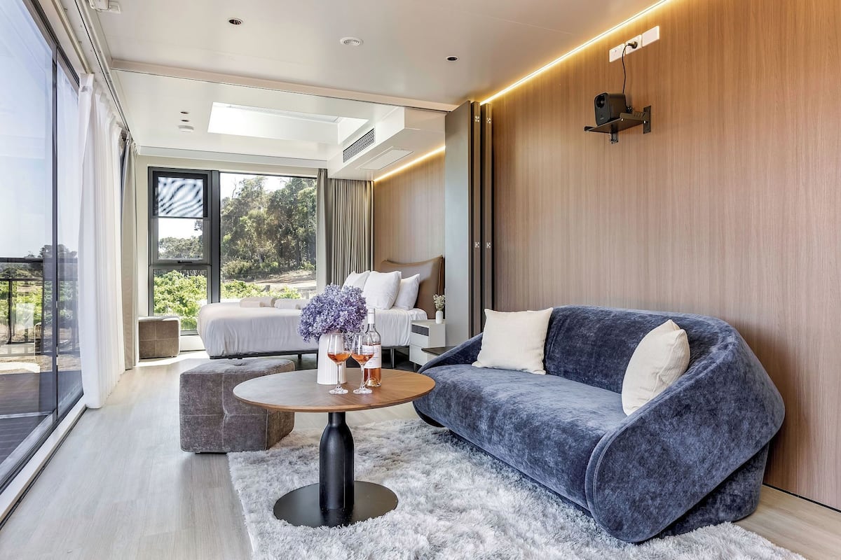 A modern, well-lit room with a large window, a cozy blue sofa, a round wooden table, and a bed with a view of greenery outside.