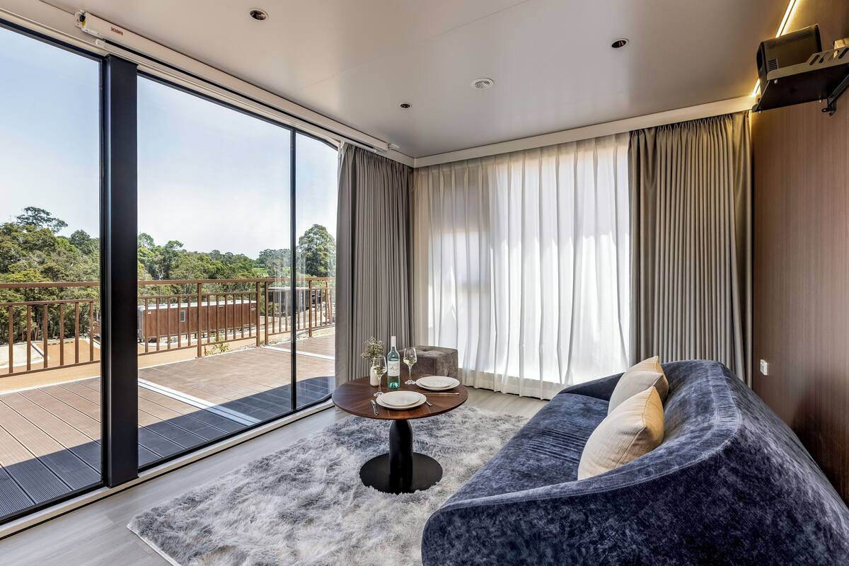 A modern living room with a large window, a round table with a rug, a sofa, and a balcony with a view of greenery.