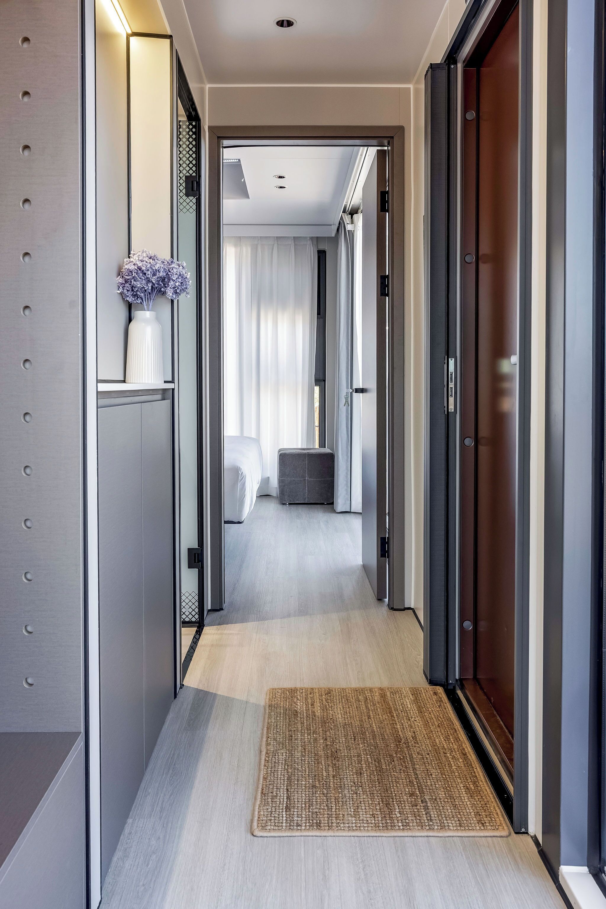 A modern hotel corridor with a neutral color scheme, a visible bed, and a small seating area.