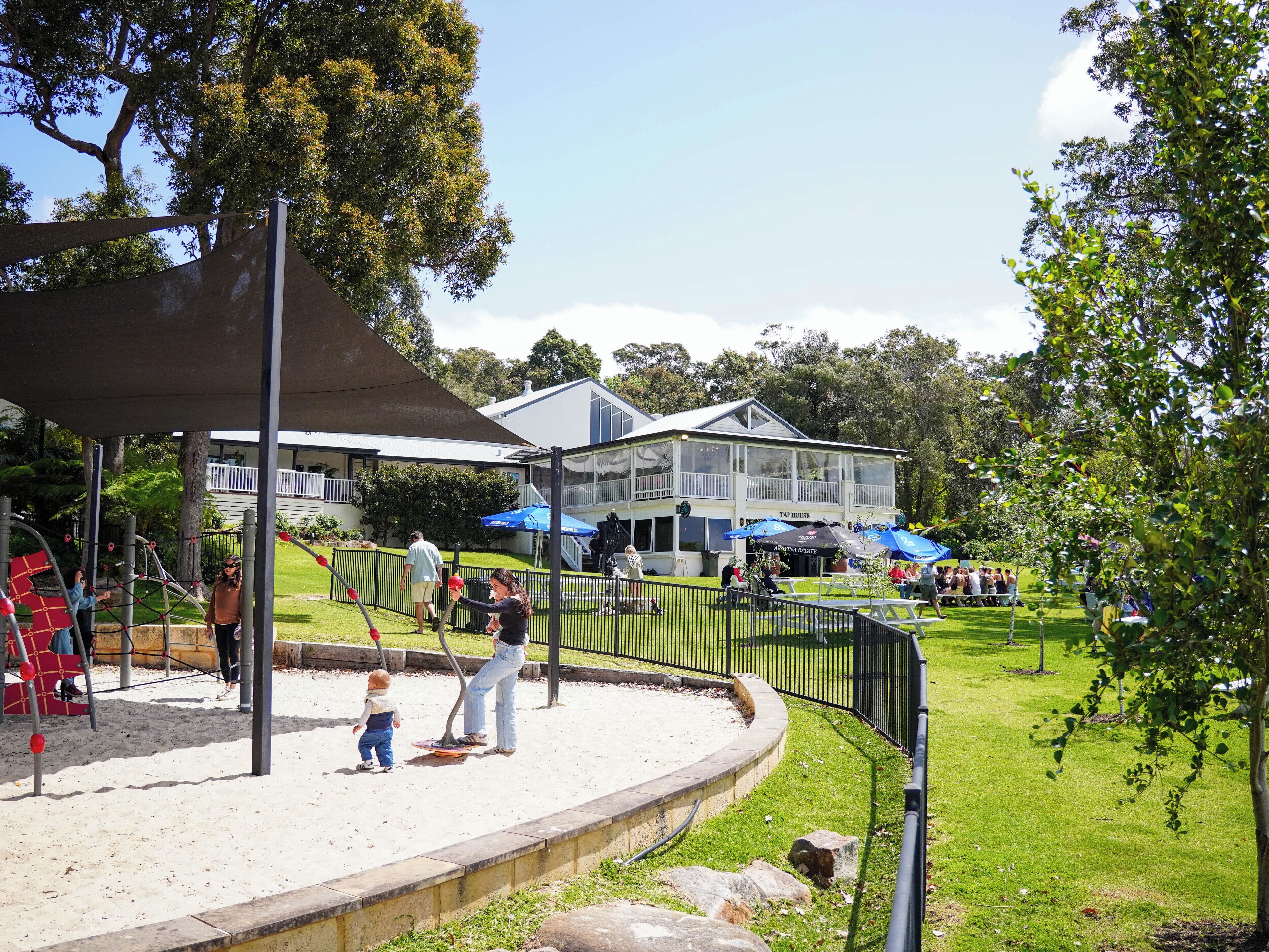 Un parque con un área de juegos, un edificio con la leyenda 'TAP HOUSE' y una zona verde con gente.