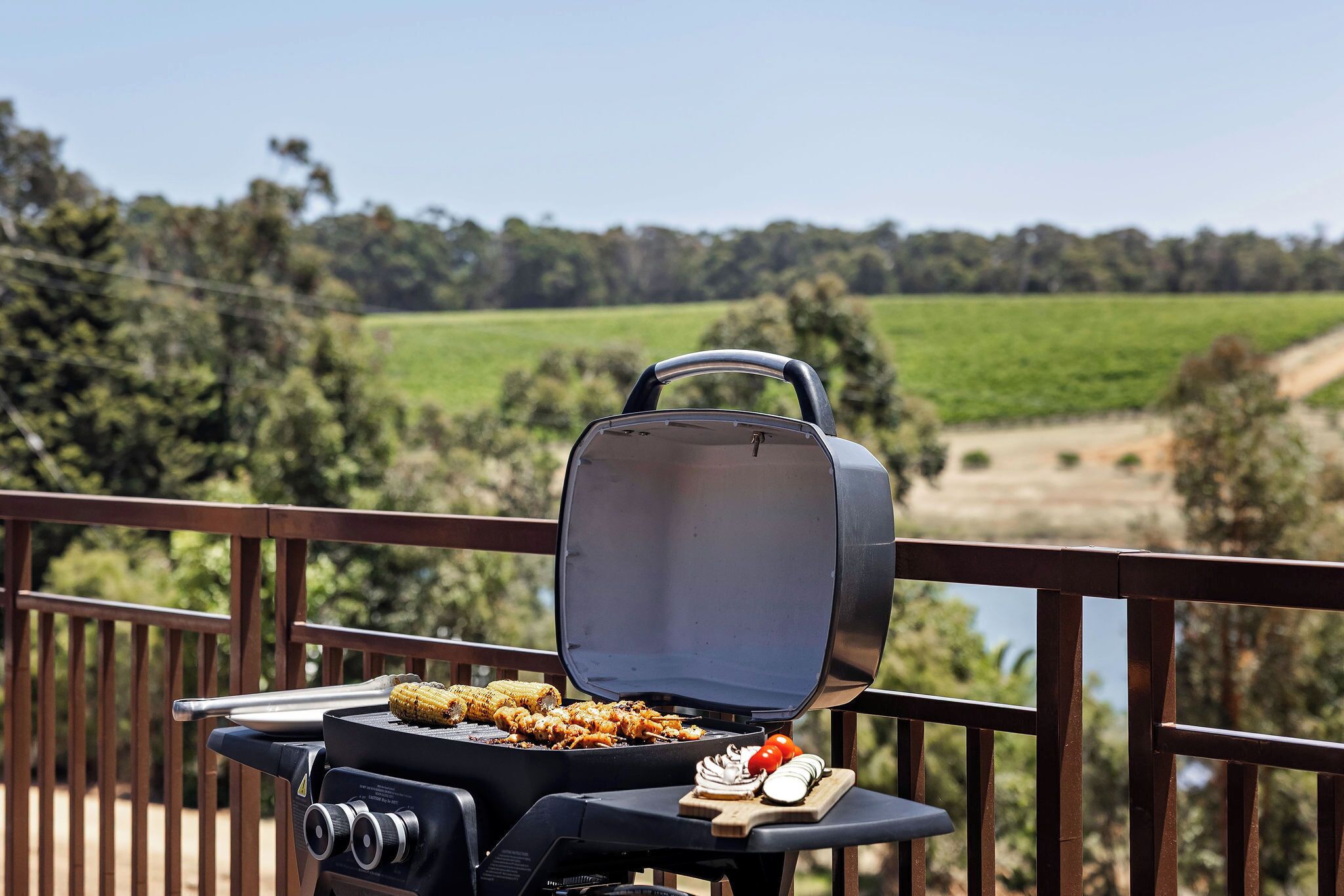 Ein Grill mit Mais und Gemüse auf einem Balkon mit Blick auf eine grüne Landschaft.