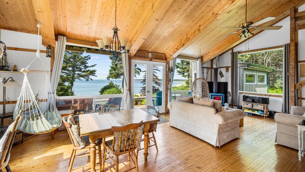 A spacious living area with a wooden ceiling, a dining table with chairs, a sofa, and a view of the sea. - Megvy: Oceanfront PEI Beach House with Gazebo, Deck & Private Beach Access (Prince Edward Island)