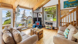 A cozy cabin interior with a wooden ceiling, a sofa, a wooden chest, a hanging chair, and a view of the ocean. - Megvy: Oceanfront PEI Beach House with Gazebo, Deck & Private Beach Access (Prince Edward Island)