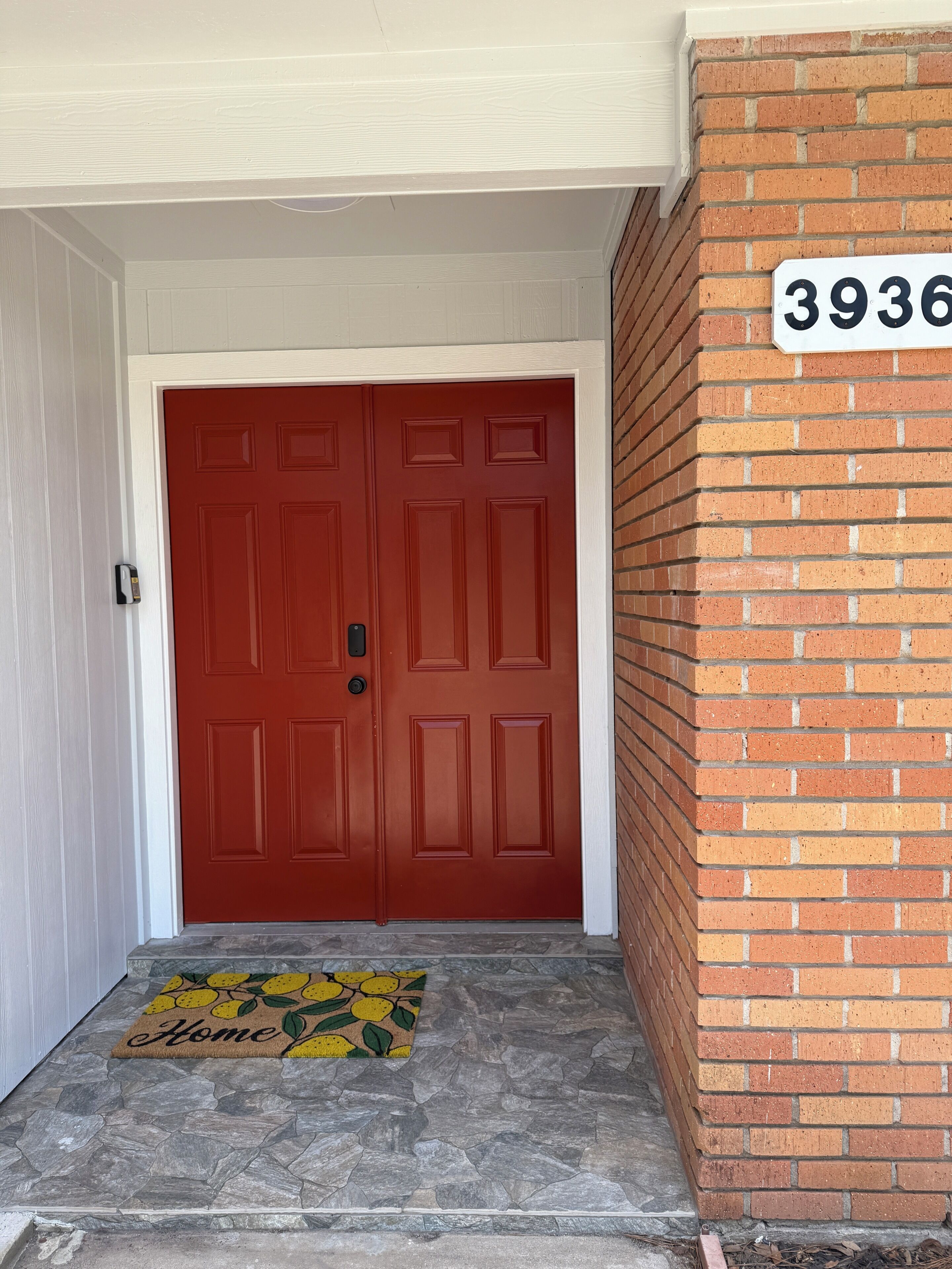 A red double door with a mailbox on the left, a stone-tiled entrance, a yellow doormat with 'Home' on it, and a brick wall with the number '3936' above the door.