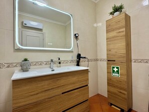 A bathroom with a wooden vanity, a large mirror, a hairdryer, and a first aid sign.