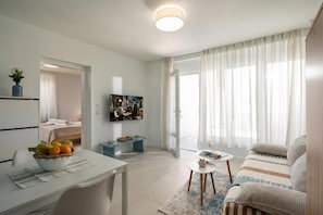 A modern living room with a sofa, a dining table with a bowl of fruit, a TV, and a large window with curtains.
