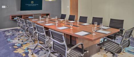 A conference room with a long table, 10 chairs, and a large screen displaying the hotel name.