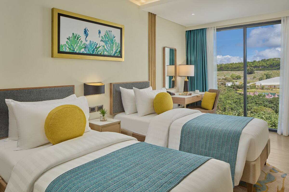 A hotel room with two beds, a desk, a chair, a large window with a view of greenery, and a framed seahorse artwork on the wall.