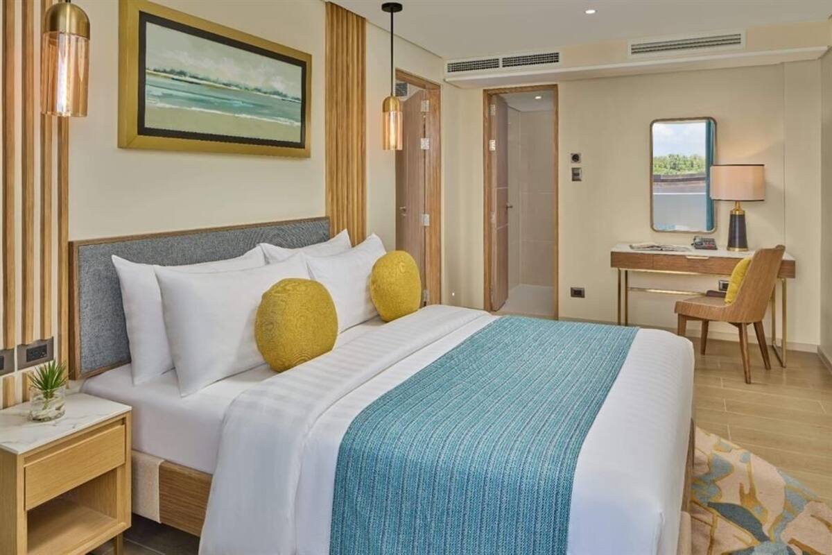 A hotel room with a bed, bedside table, lamp, chair, desk, and a painting on the wall.
