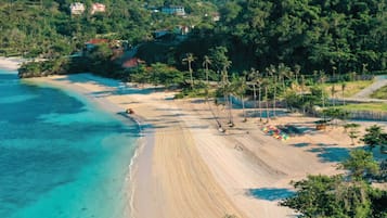A beach with clear turquoise waters, a sandy shore, and a backdrop of lush green hills with buildings.