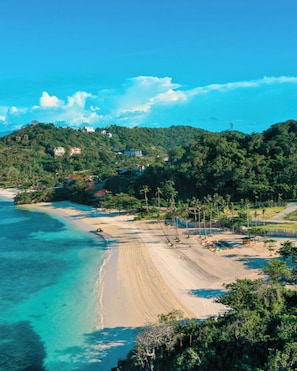 Una playa con aguas turquesas cristalinas, una orilla arenosa y colinas verdes exuberantes con edificios al fondo.