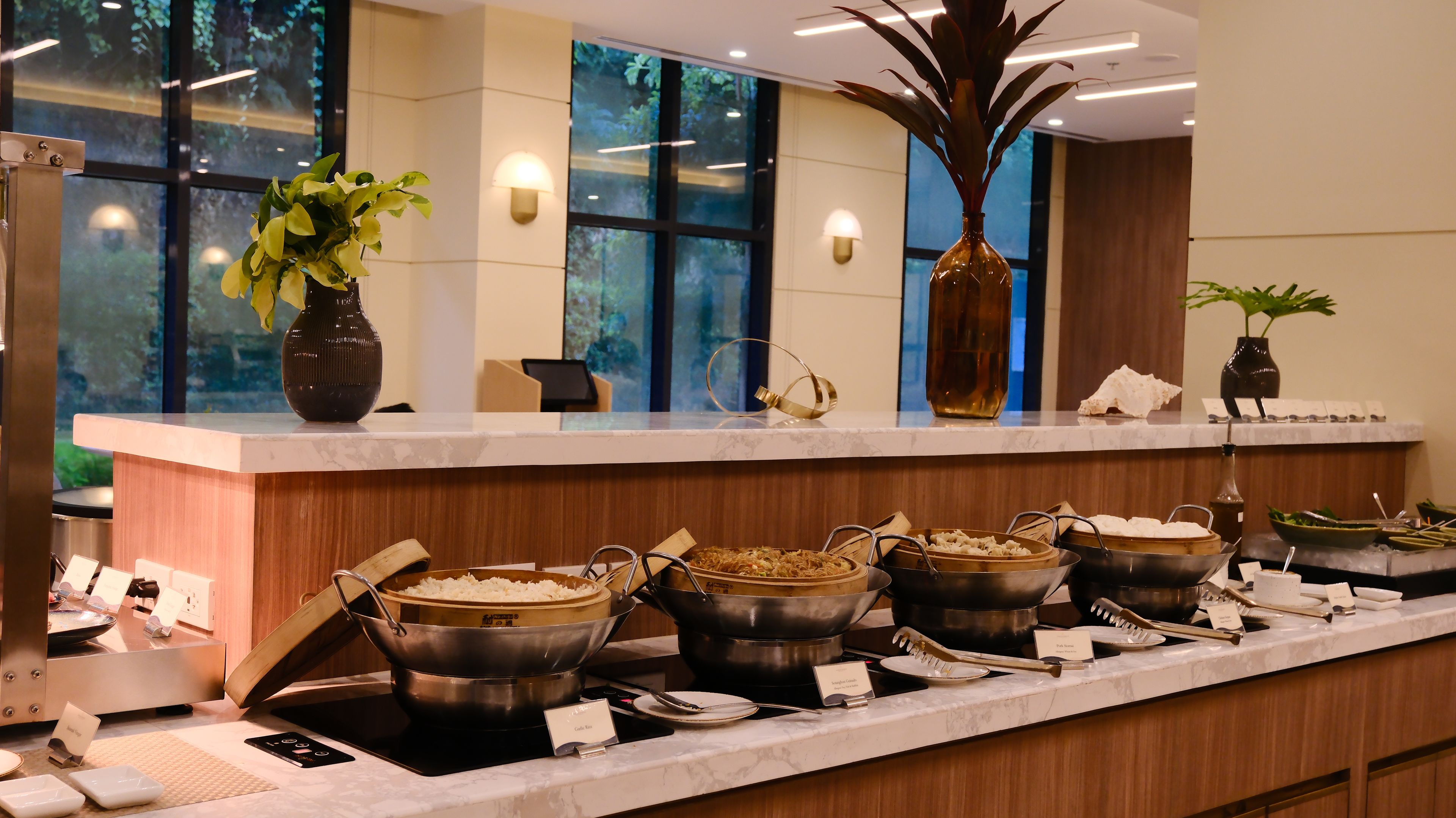 A buffet setup with various dishes and utensils, a vase with a plant, and a window with a view of greenery.