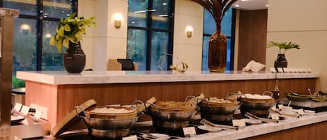 A buffet setup with various dishes and utensils, a vase with a plant, and a window with a view of greenery.