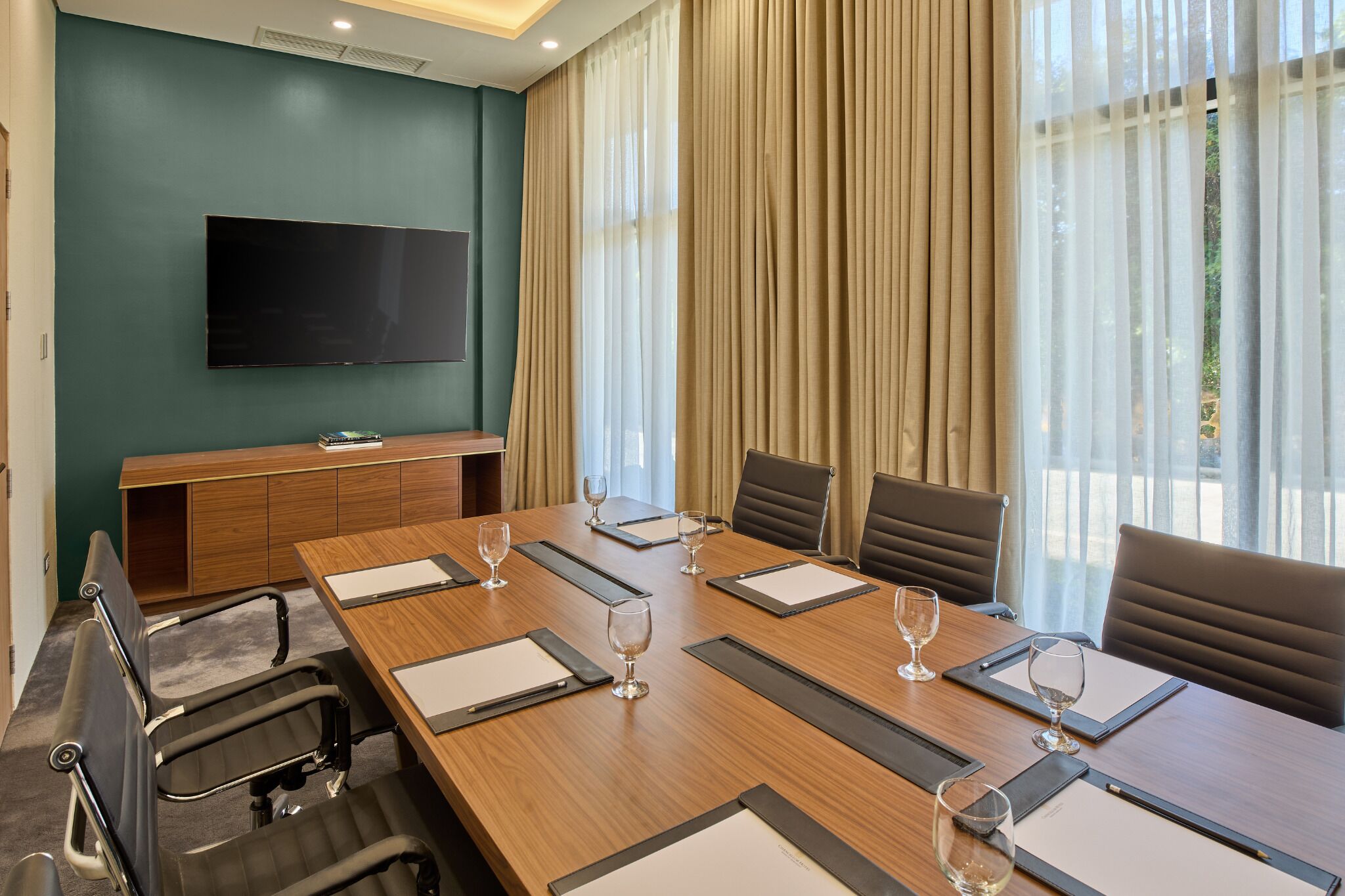 A conference room with a long table, chairs, a TV mounted on the wall, and large windows with curtains.