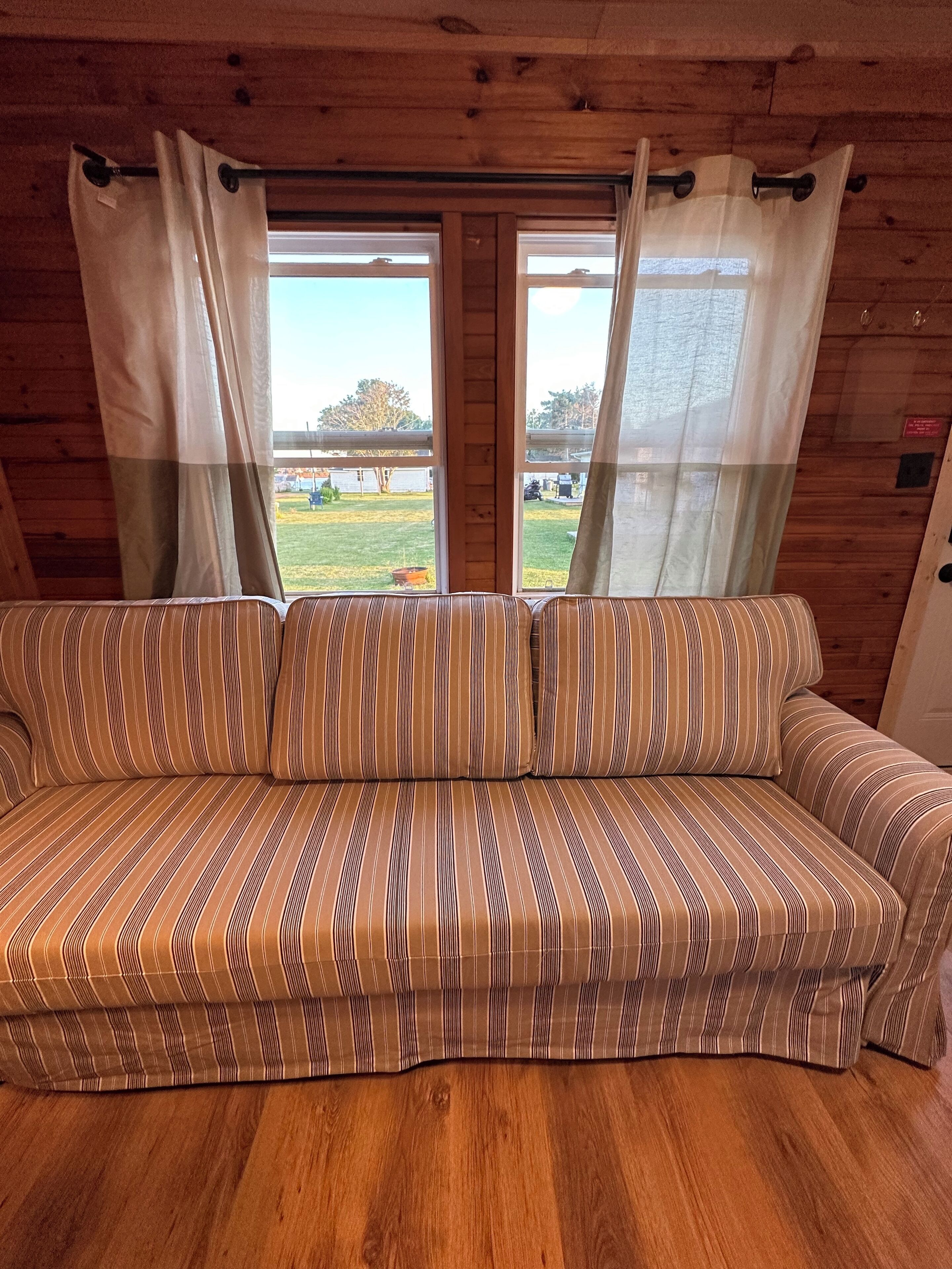 L’intérieur d’un chalet en bois, avec un canapé à rayures, deux fenêtres avec des rideaux et une vue sur une zone herbeuse.