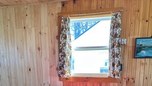 Una habitación con paneles de madera, una mesa redonda cubierta con mantel a cuadros, sillas rojas y una ventana con cortinas florales.