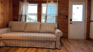 Interior de una cabaña de madera con un sofá a rayas, dos ventanas con cortinas, una puerta y piso de madera.