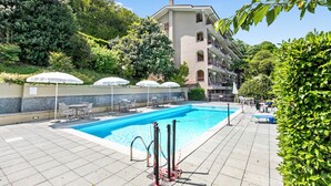 A swimming pool with lounge chairs and umbrellas, surrounded by greenery and a building with balconies.