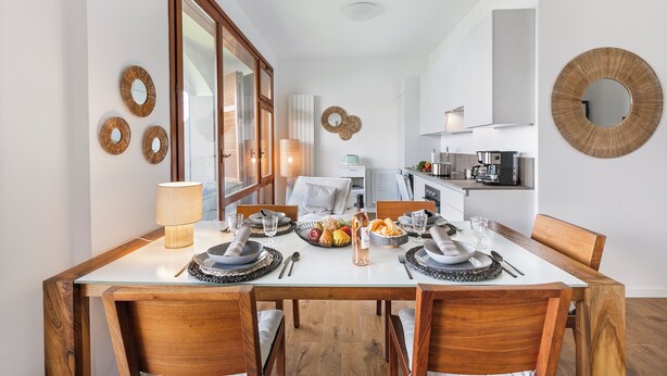 A dining area with a wooden table set for four, surrounded by chairs. A kitchen is visible in the background.
