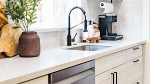 Une cuisine moderne avec des meubles blancs, un lave-vaisselle en acier inoxydable, une machine à café et une fenêtre équipée de stores.