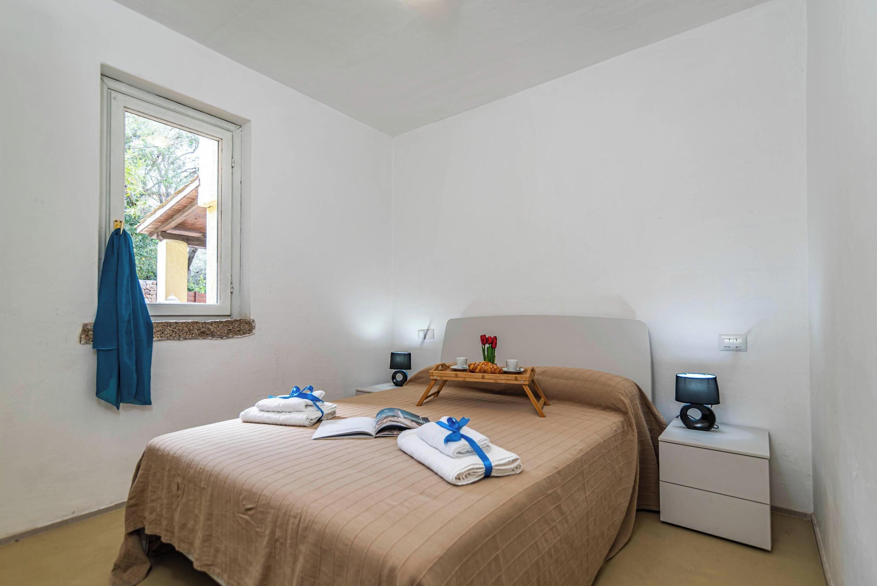 A neatly made bed with towels and a tray of breakfast items, a bedside table with a lamp, and a window with a towel hanging outside.